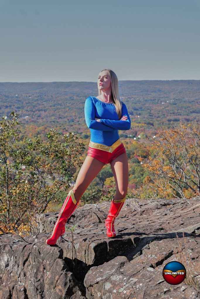 Mandii Monarch as UltraGirl, arms folded, on a rocky outcrop above autumn hills